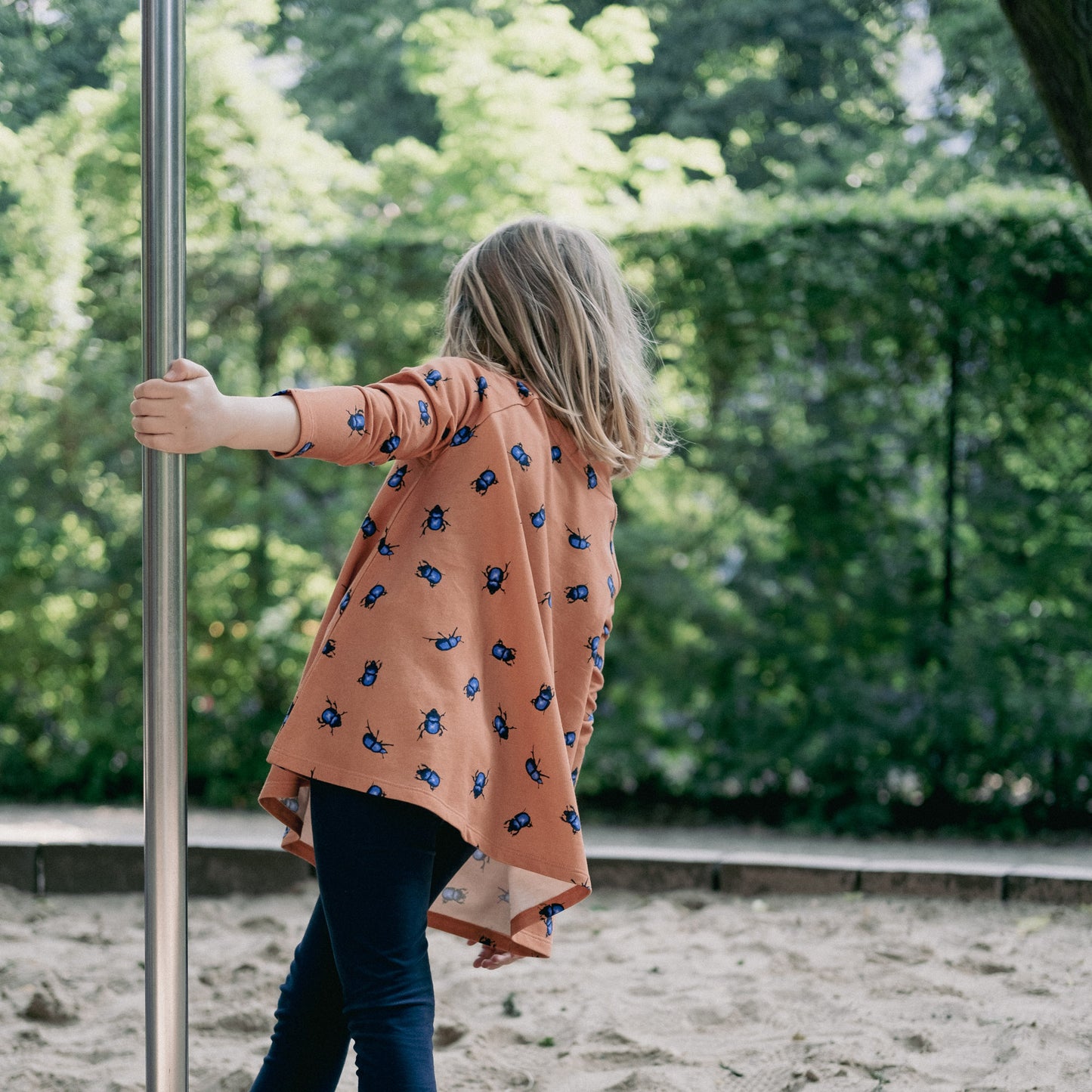 Girl in a playground with a orange tunic dress with blue beetles on.