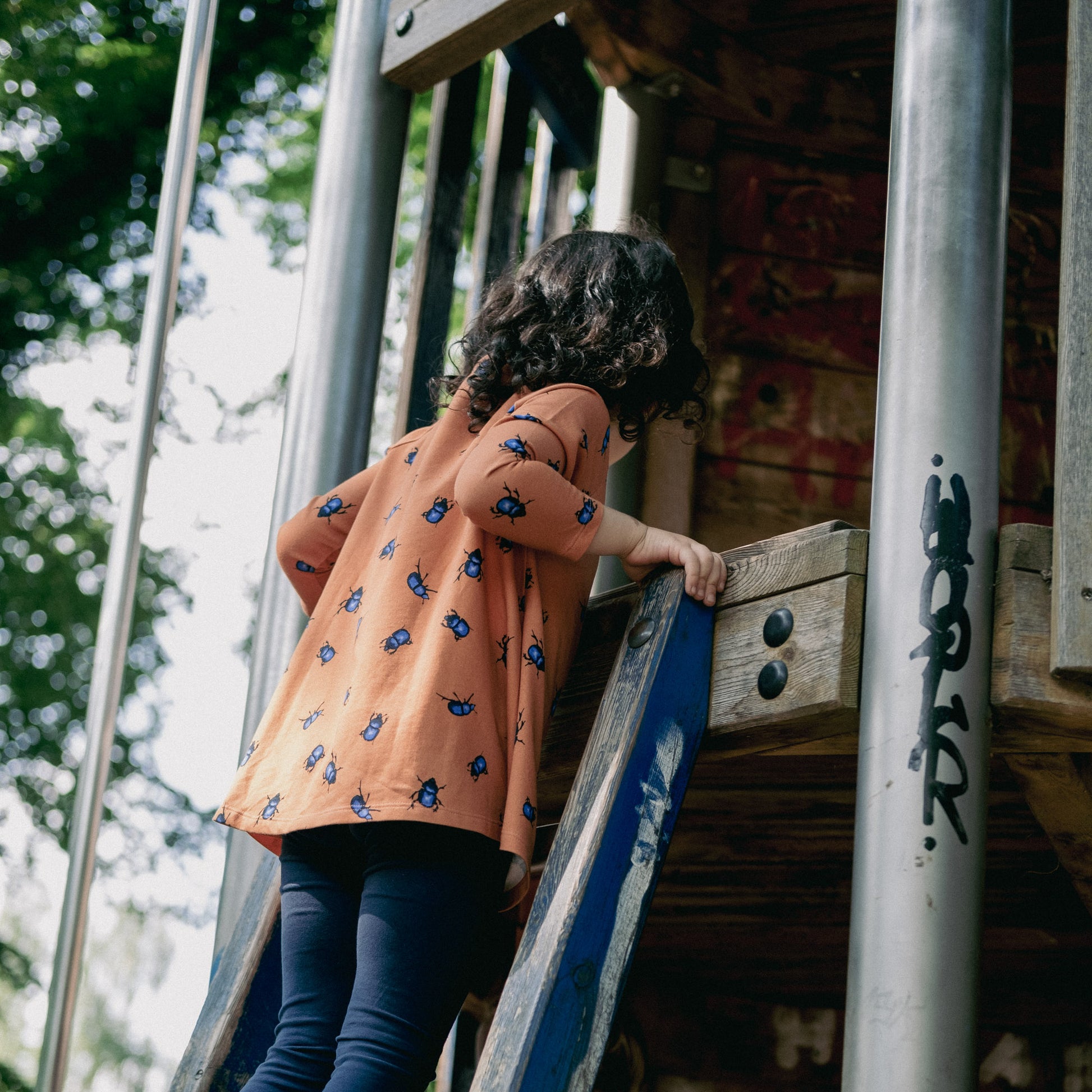 Girl climbing in a climbing frame with a orange tunic with beetles on.