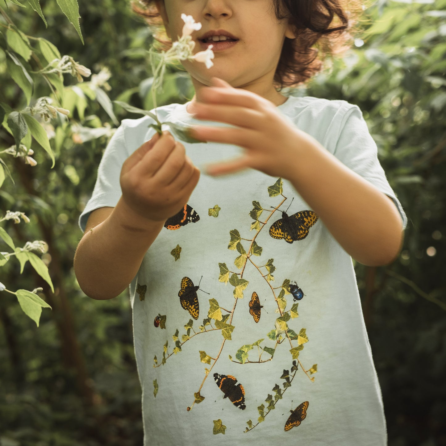 Girl playing in bushes summertime holding a flower. Wearing a green t-shirt with butterflies and ivy.  