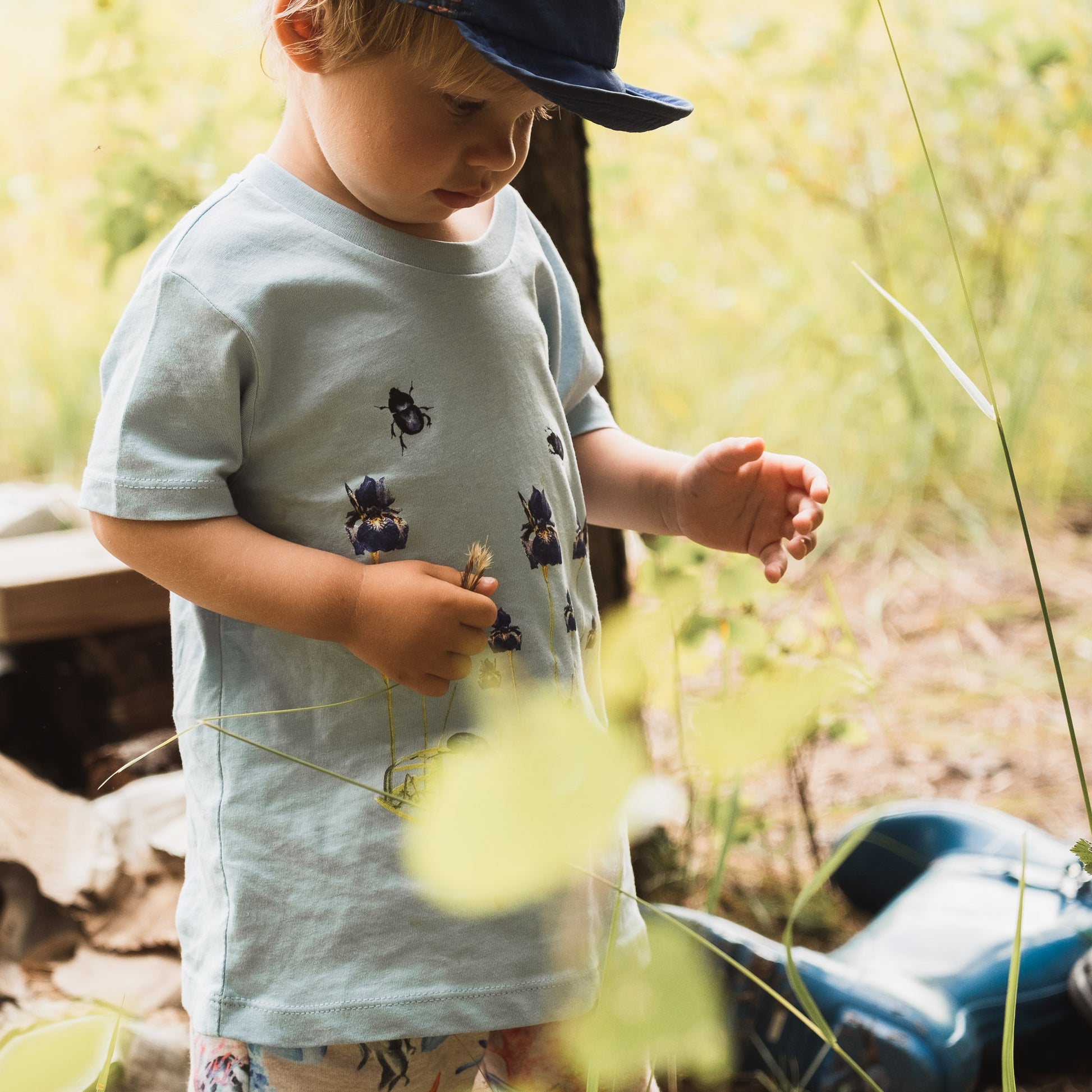 Toddler feeling grass during the Swedish summer. Wearing a blue cap, blue t-shirt with irises and beetles and shorts with fish. In the background there are blue boots and a pile of logs.