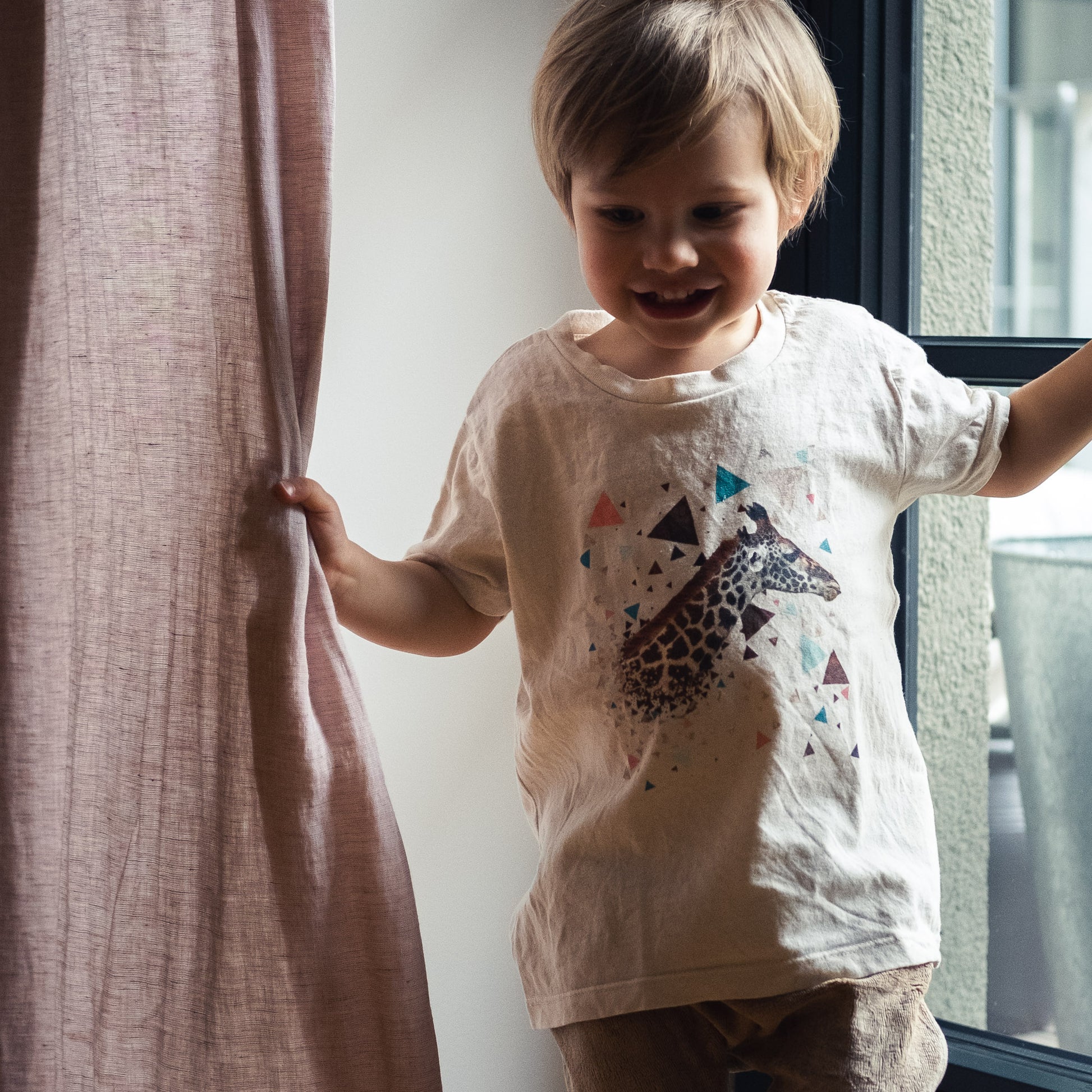 Toddler wearing a natural coloured t-shirt with a giraffe and colourful triangles. Playing with pink curtains.